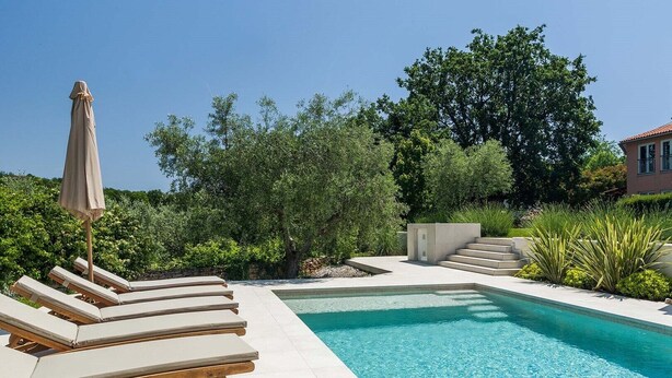 A pool area with lounge chairs and a covered umbrella, surrounded by greenery and a building in the background.