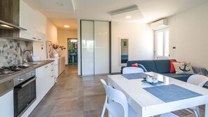 A modern kitchen with a gas stove, white cabinets, and a tiled backsplash. There is a dining area with a white table and chairs, and a sofa with cushions.