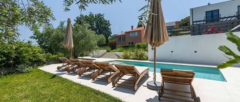 Un espace aménagé au bord de la piscine, avec des chaises longues et des parasols, une piscine à l’eau d’un bleu limpide et un bâtiment moderne en arrière-plan.