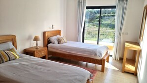 Ett sovrum med två sängar, ett sängbord, ett litet bord vid sängen och utsikt över en trädgård genom ett stort fönster.
