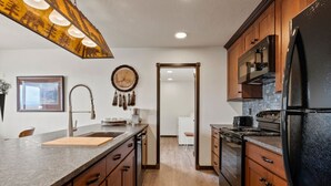 A kitchen with wooden cabinets, a granite countertop, a black refrigerator, and a hanging light fixture. - Newly Built Rustic Cabin w/ Mountain & Pond Views (Kalispell)