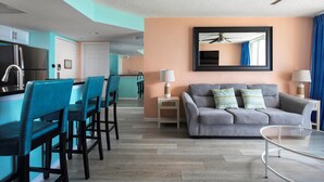 Una sala moderna con un sofá gris, sillas color turquesa, una mesa de centro de vidrio y una zona de cocina con refrigerador y armarios.