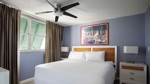Un dormitorio con cama, mesitas de noche, ventilador de techo, ventana con cortinas y un cuadro en la pared.