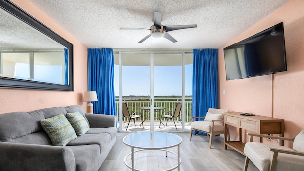 Habitación de hotel con sofá, una mesa redonda de vidrio, televisión, balcón con cortinas azules y ventilador de techo.