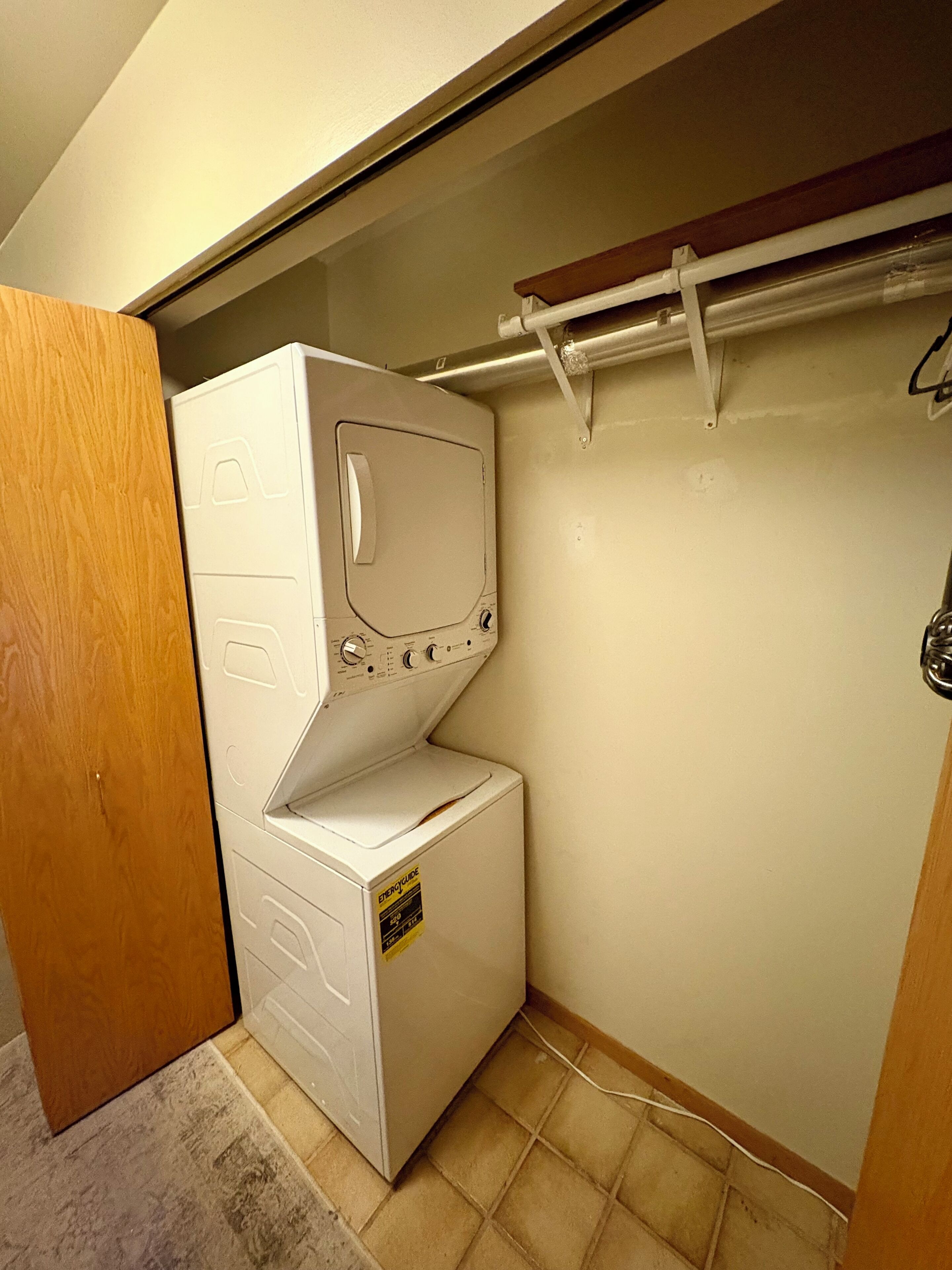 Une petite buanderie équipée d’un lave-linge et d’un sèche-linge superposés, d’une porte en bois et d’un sol carrelé.