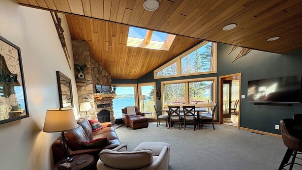 Amplia sala de estar con chimenea de piedra, techo de madera y grandes ventanas con vistas panorámicas.