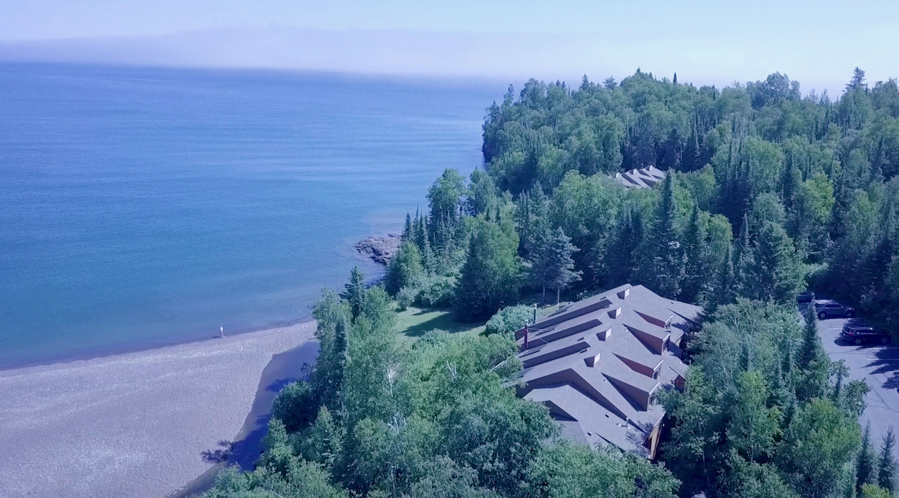 An aerial view of a coastal property with a beach, dense forest, and a house with a unique roof design.