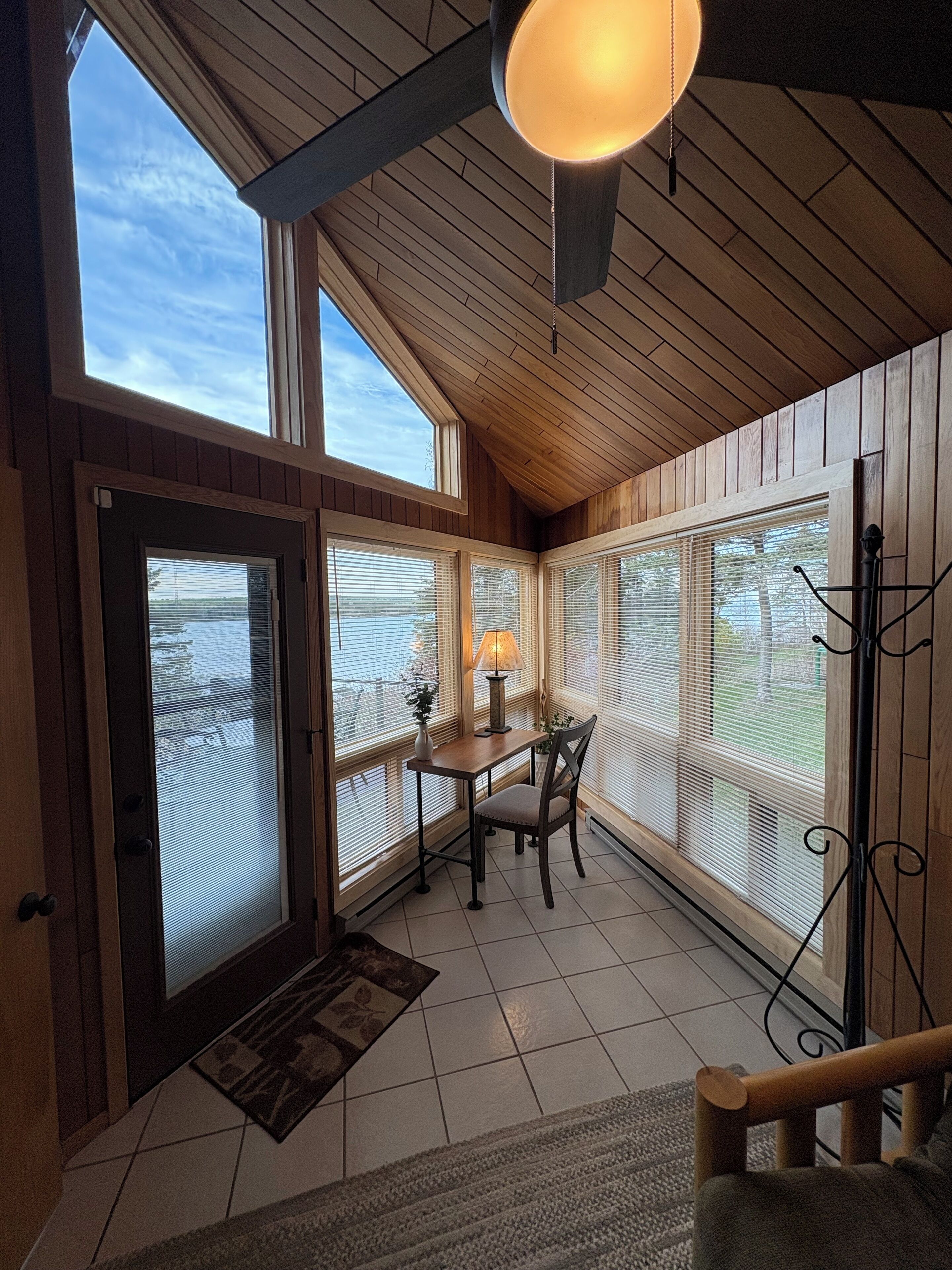 A cozy room with wooden walls and a sloped ceiling, featuring a table with chairs, a lamp, and a view of the outdoors through large windows.