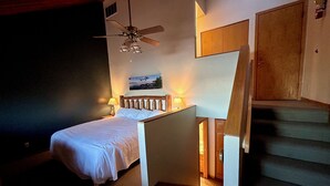 Un dormitorio con cama, ventilador de techo, un cuadro, una mesita de noche y una escalera.