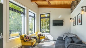 A modern living room with large windows, a yellow armchair, a gray sofa, and a flat-screen TV mounted on the wall.