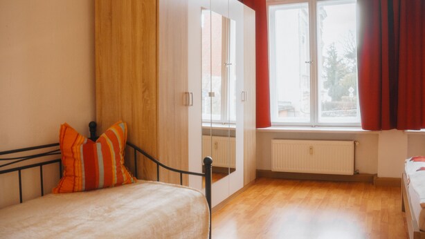 Een slaapkamer met een bed, een raam met gordijnen, een radiator en houten vloeren.