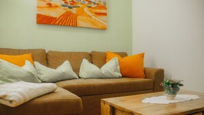 Een woonkamer met een bruine bank, oranje en witte kussens, een houten salontafel en een schilderij aan de muur.