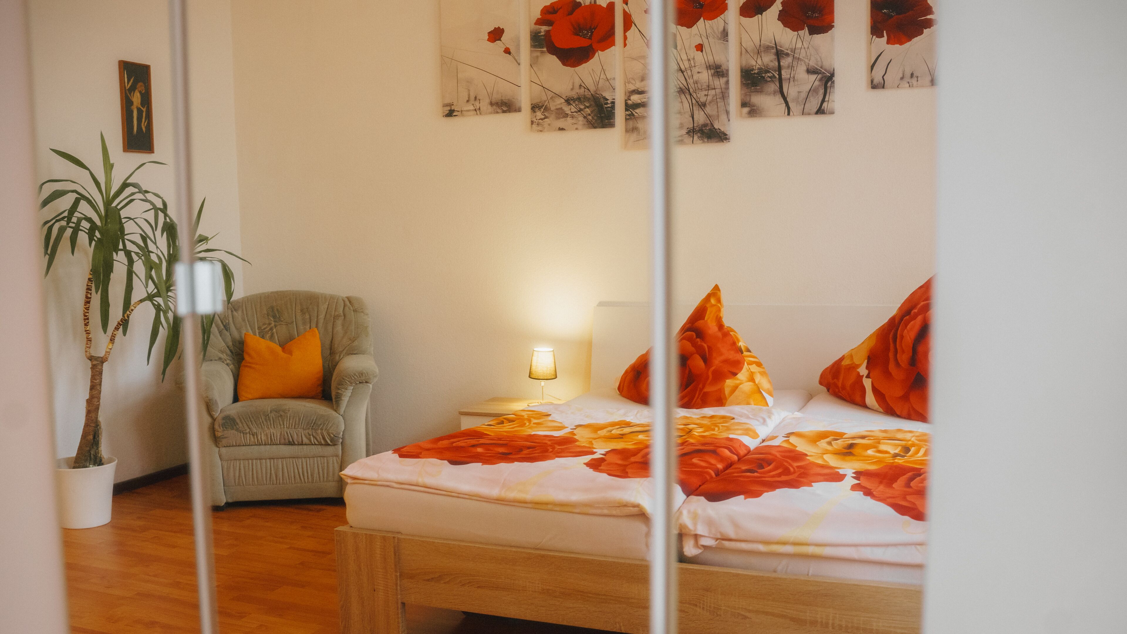Un dormitorio con cama, una silla, una mesita de noche con lámpara, una pared decorada con una obra de arte y una planta en maceta.