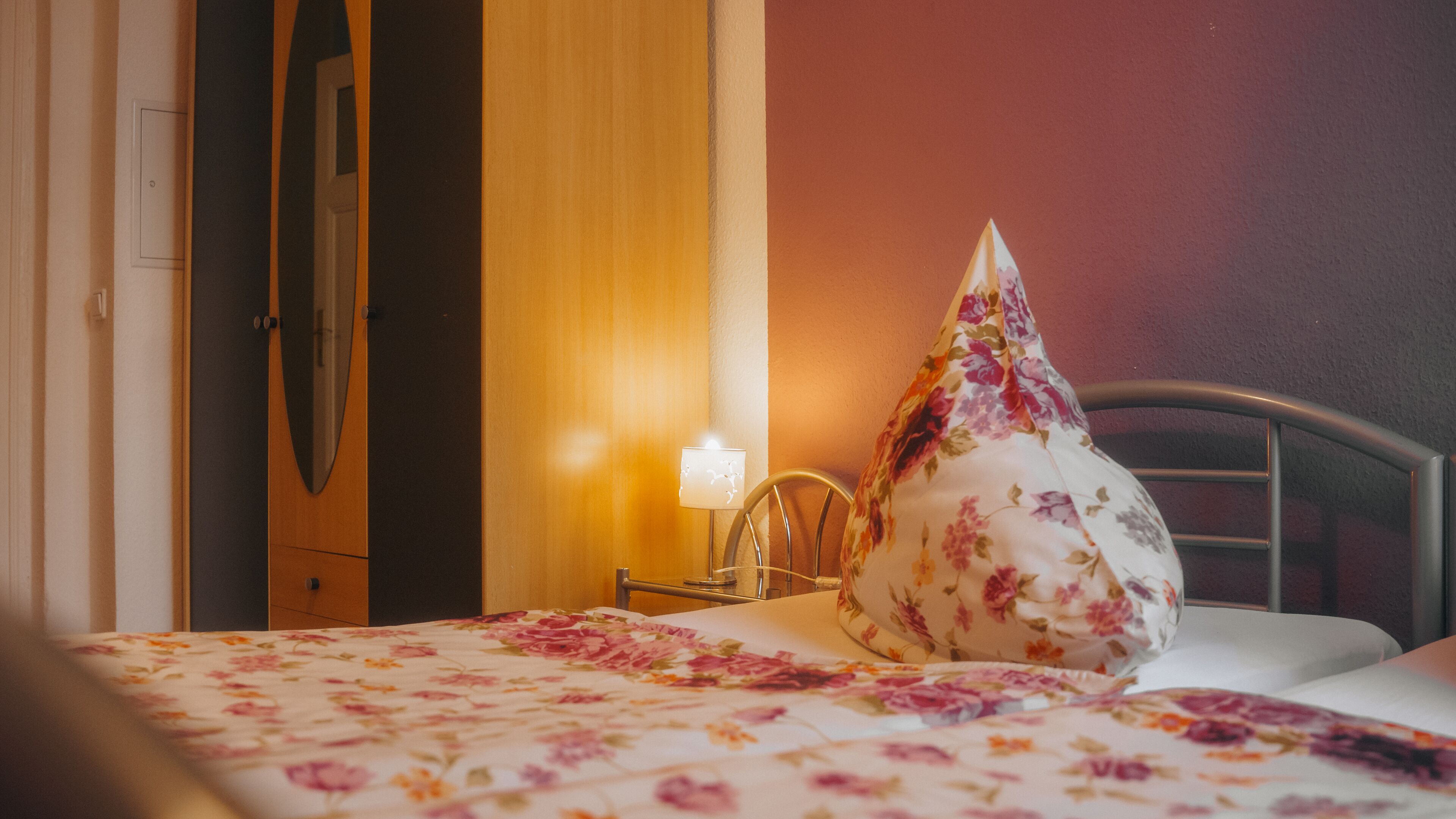 A hotel room with a bed, a nightstand, a lamp, and a floral-patterned pillow.