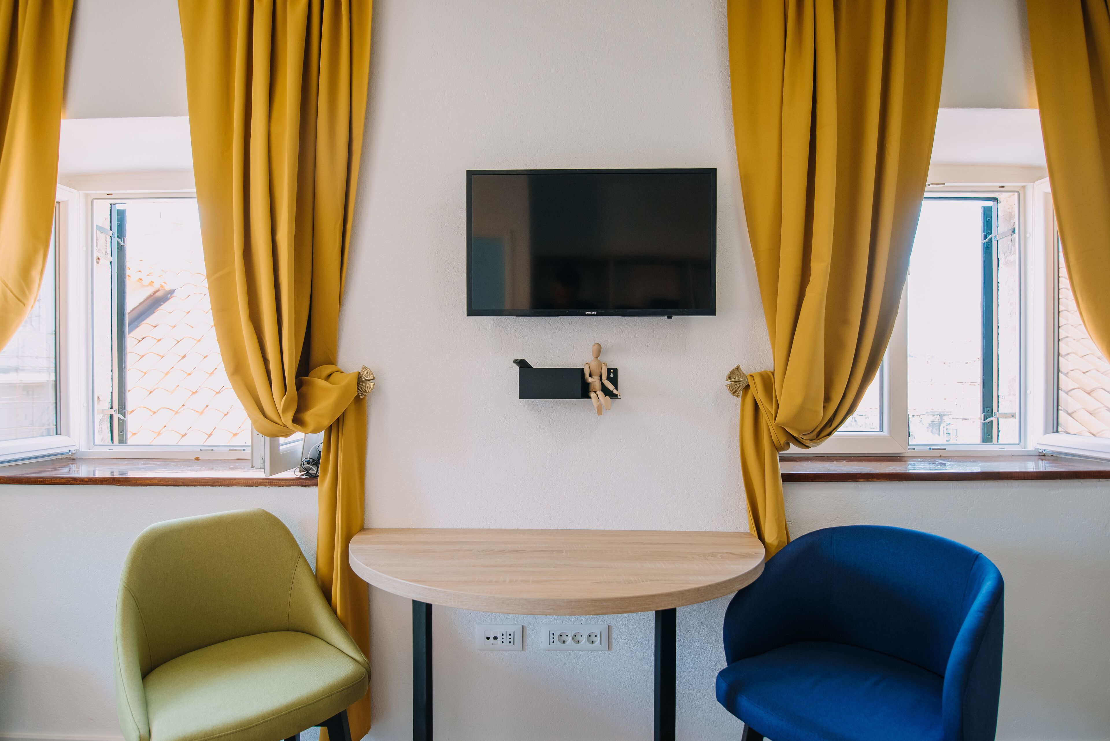 A room with a TV mounted on the wall, a small table, two chairs, and yellow curtains.