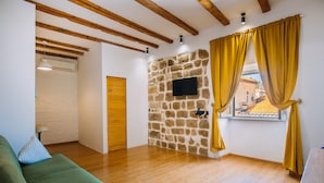 A room with a stone wall, wooden beams, a green sofa, and a window with yellow curtains. - Art Deco Cathedral Apartments by DuHomes (Dubrovnik)