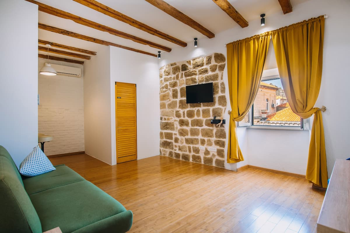 Una habitación con pared de piedra, vigas de madera, un sofá verde y una ventana con cortinas amarillas.