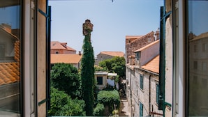 A view through a window of a narrow street with buildings, a tall tree, and a small structure with a white cloth hanging. - Art Deco Cathedral Apartments by DuHomes (Dubrovnik)