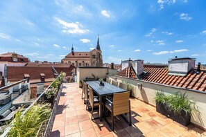 A rooftop terrace with a dining table and chairs, potted plants, and a view of a historic building with a spire.