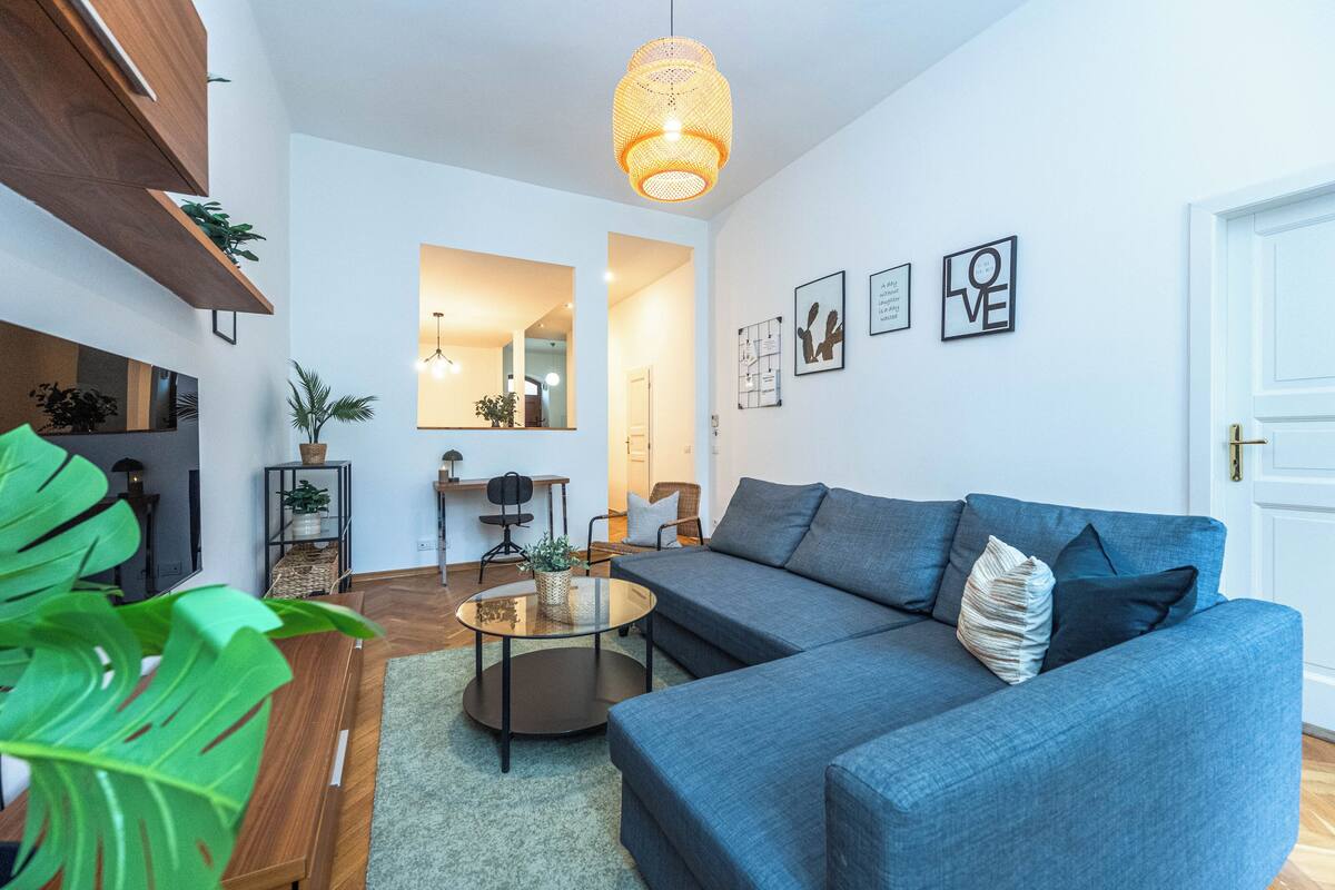 A modern living room with a grey sofa, a round coffee table, and a hanging light fixture. There are framed pictures on the wall and a door to the right.