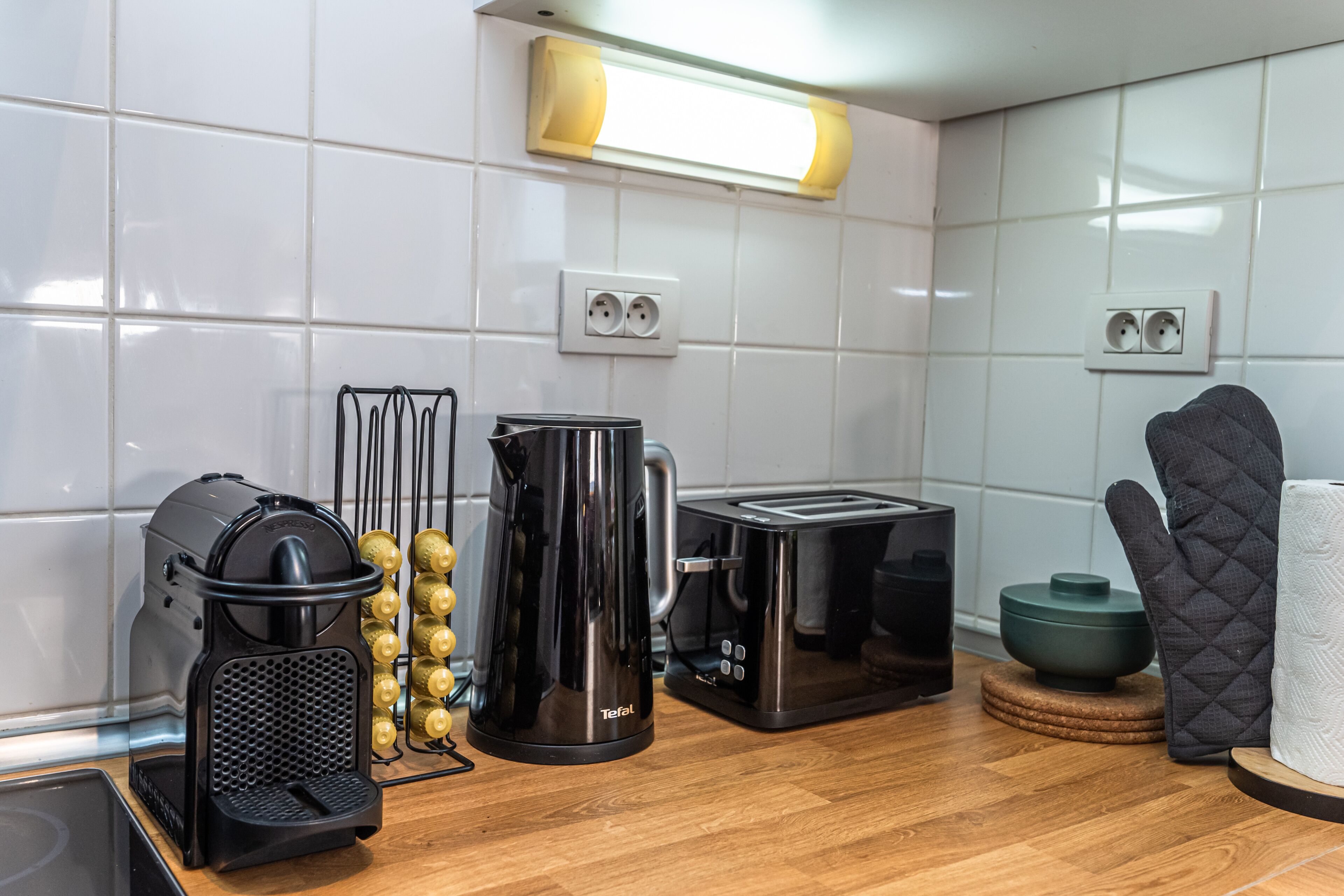A modern kitchen with white tiled walls, wooden flooring, a coffee maker, a toaster, a kettle, and a kettle holder.