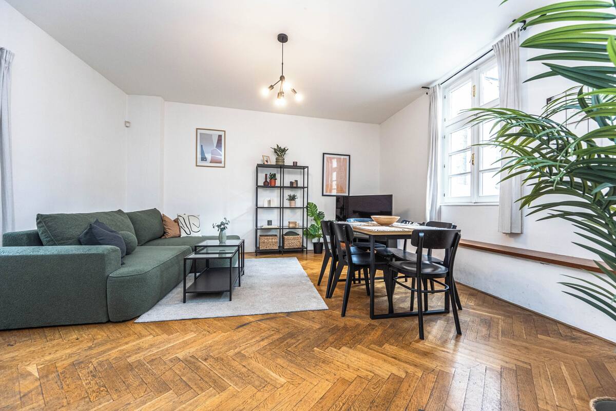 A living room with a green sofa, a dining table with chairs, a bookshelf, and a large window with curtains.