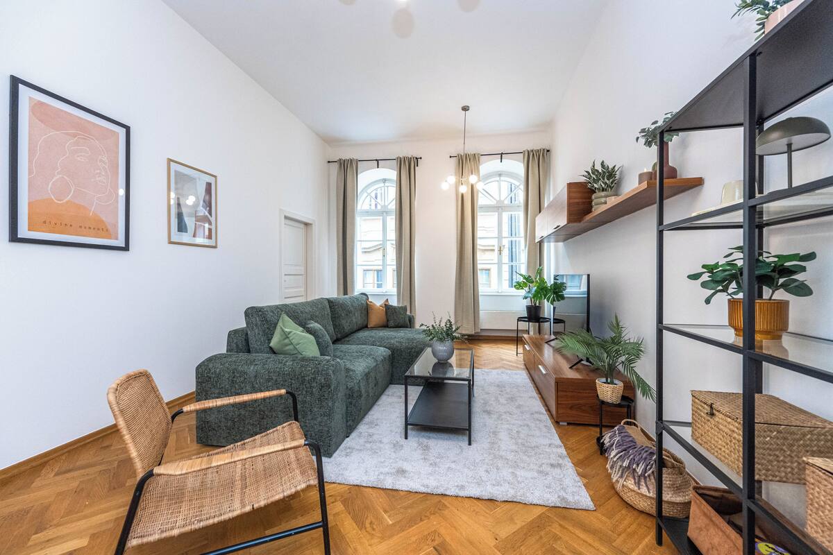 A modern living room with a green sofa, a wicker chair, a coffee table, and a bookshelf filled with plants.