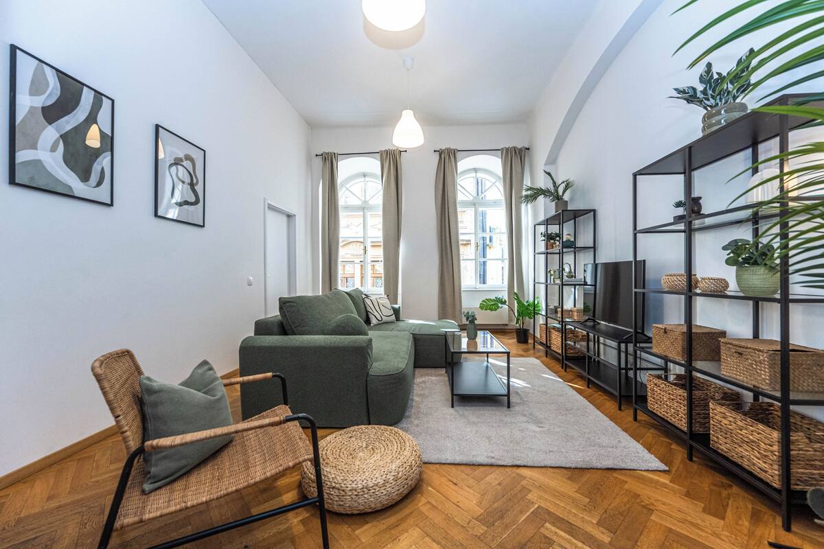 A modern living room with a sofa, armchair, and coffee table, a bookshelf, and large windows.