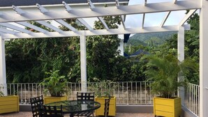 Une terrasse couverte avec une table ronde et des chaises, entourée de plantes en pot et d’une balustrade blanche.