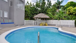 Une piscine entourée d’une clôture blanche, un abri au toit de chaume et une végétation luxuriante en arrière-plan.