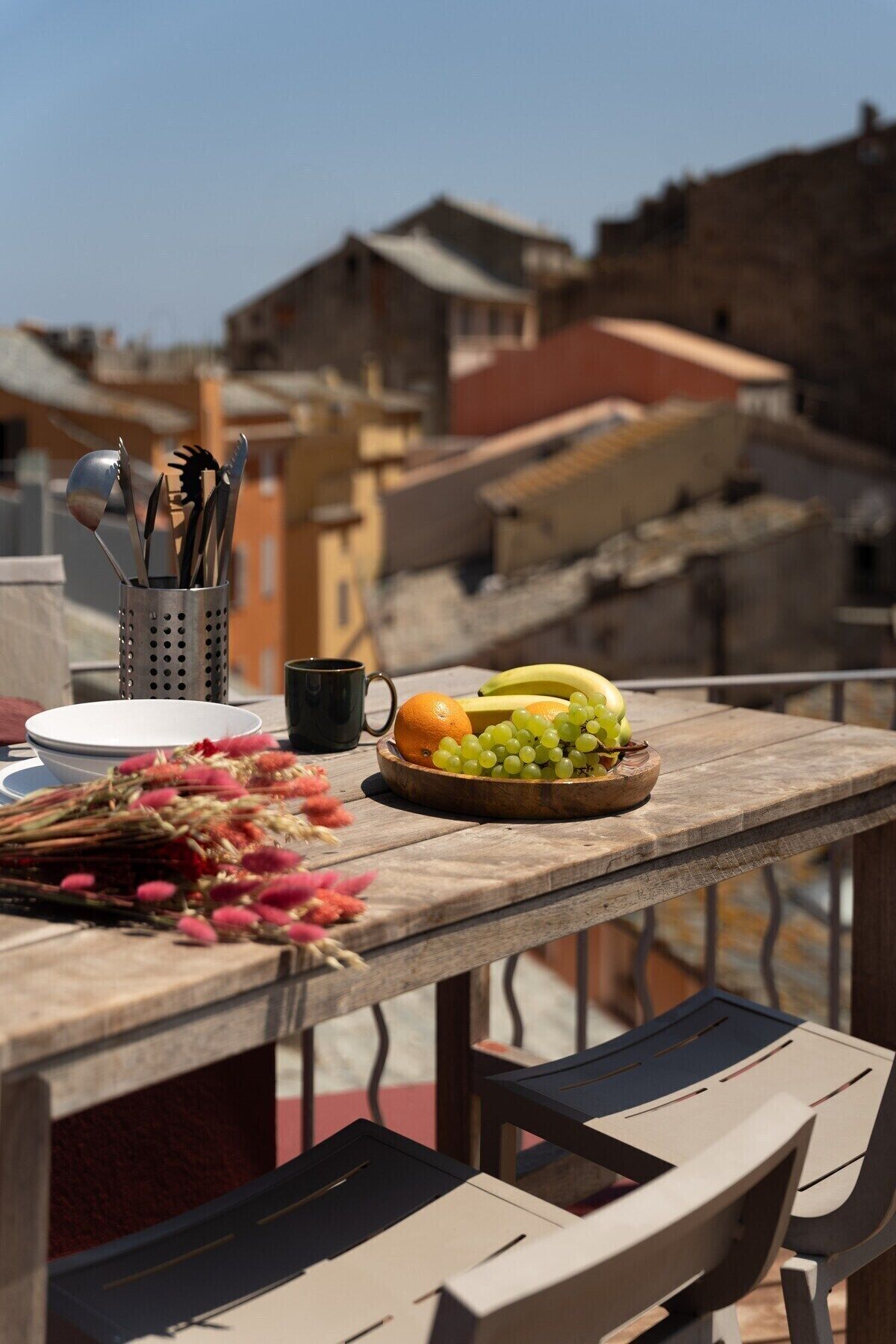 Une table en bois sur laquelle se trouve un panier de fruits contenant des raisins, des bananes et des oranges, un porte-ustensiles en métal, une tasse noire et un bouquet de fleurs, le tout placé sur un balcon donnant sur des immeubles.