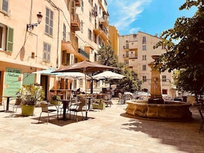 Terrassa exterior d'un cafè amb taules i caires, envoltada d'edificis amb balcons i una fontania.