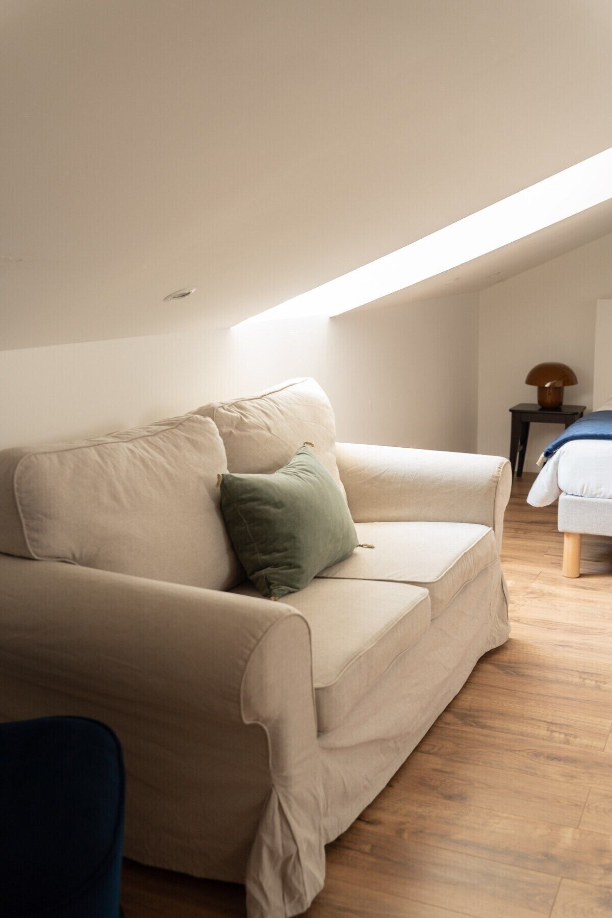 Een moderne, minimalistische woonkamer met een beige bank, een groen kussen en een kleine tafel met een lamp.