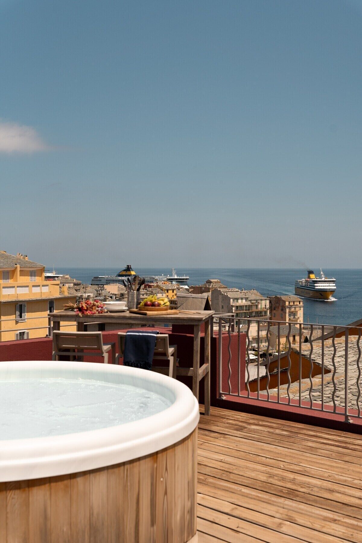 Una terraza de madera con un jacuzzi, una mesa con fruta y vistas al mar y barcos.