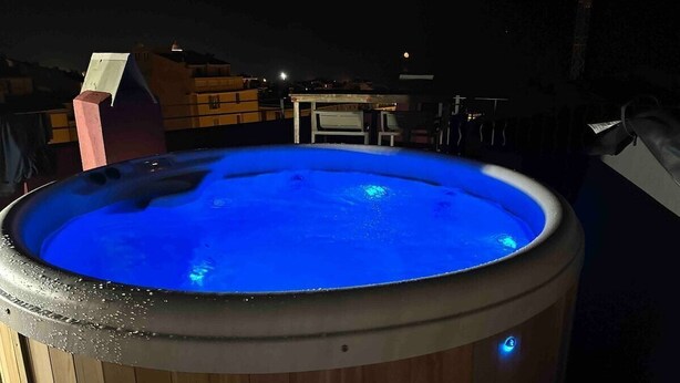 Un jacuzzi iluminado con luz azul, rodeado de una terraza de madera y una mesita.