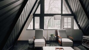 A room with a sloped ceiling, large windows, two chairs, a small table, and a lamp. - Modern A-Frame | Mountain Views | Evergreen (Evergreen)