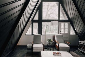 A room with a sloped ceiling, large windows, two chairs, a small table, and a lamp. - Modern A-Frame | Mountain Views | Evergreen (Evergreen)