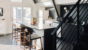 A modern kitchen with a large island, a dining area with chairs, a staircase, and a view of the outdoors. - Modern A-Frame | Mountain Views | Evergreen (Evergreen)