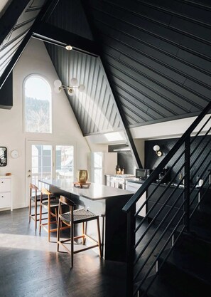 A modern kitchen with a large island, a dining area with chairs, a staircase, and a view of the outdoors. - Modern A-Frame | Mountain Views | Evergreen (Evergreen)