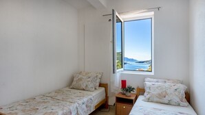 Een kleine kamer met twee eenpersoonsbedden, een raam met uitzicht op zee en een tafeltje met een rode kaars en een plant.