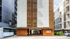 Un edificio moderno con entrada central, listones verticales de madera y un letrero que dice "D'YA HOTEL SRINAKARIN".