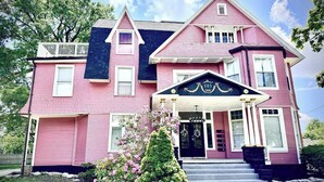 Ein pinkfarbenes, zweistöckiges Haus mit einer Veranda, einer schwarzen Tür und einem Schild mit der Aufschrift '101' über dem Eingang.