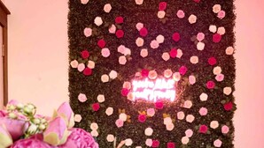 Ein Zimmer mit einer rosenbedeckten Wand und einem Neonzeichen. Ein Stuhl mit einem rosa Kissen und ein Tisch mit einem Blumenstrauß stehen dort.