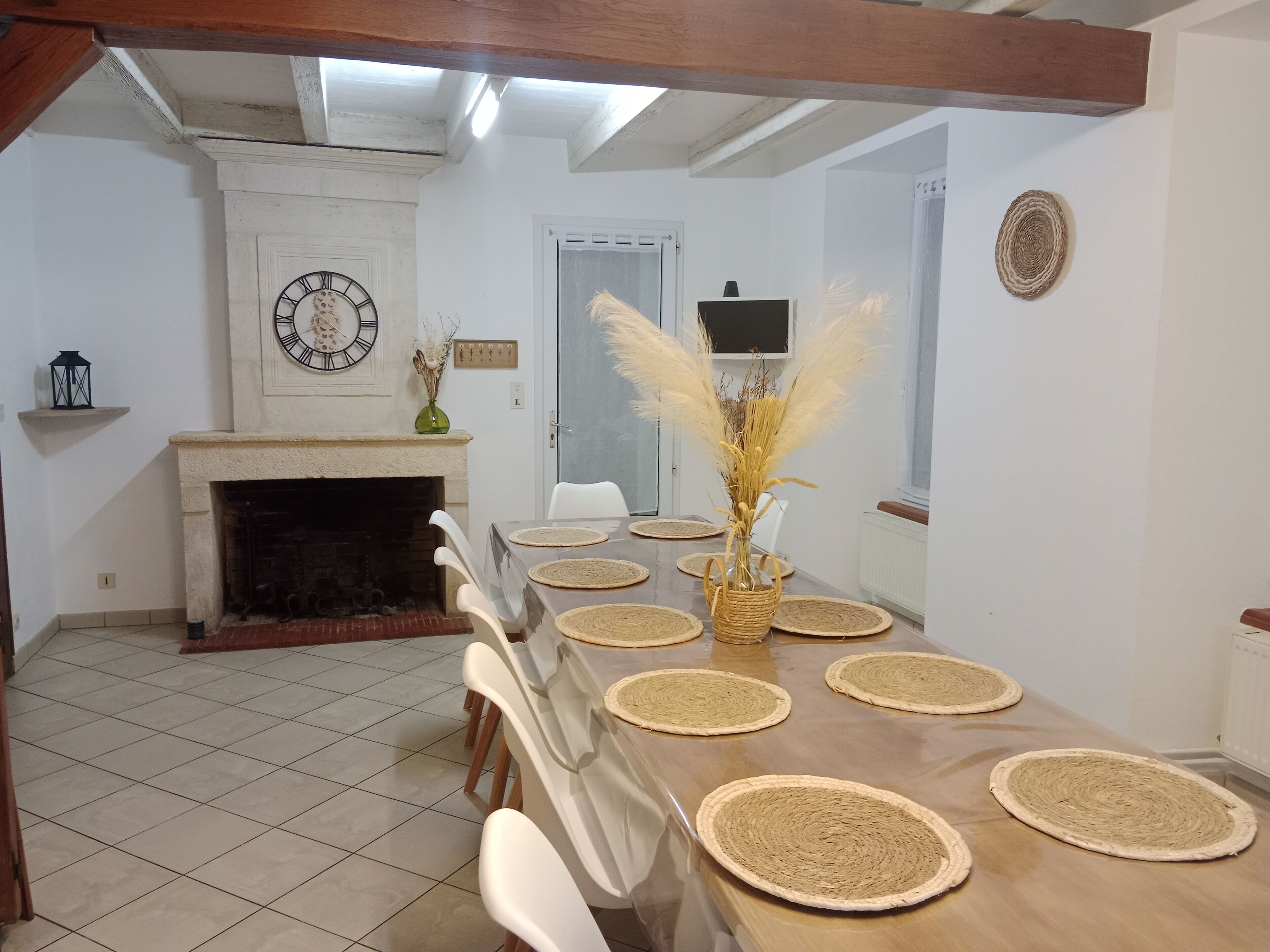 A dining room with a long table set with placemats, chairs, a fireplace, a clock, and a vase with dried plants.