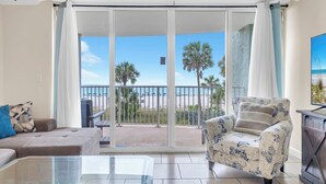 Un salon avec une table en verre, un canapé, un fauteuil à motifs et une vue sur la plage, visible à travers une porte-fenêtre coulissante.