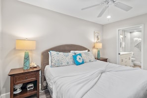 A bedroom with a bed, two bedside tables, a ceiling fan, and a bathroom visible through an open door. - Beach Service Included! | Beachfront | Steps to Gulf Pool | Windancer 101 (Miramar Beach)