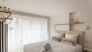Una cama bien hecha con toallas enrolladas, un cabecero, mesitas de noche y una ventana con cortinas traslúcidas.