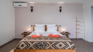 A neatly made bed with a patterned bedspread, two white pillows, and two red towels. There are two bedside tables with lamps, a wall-mounted air conditioner, and a wooden ladder against the wall.