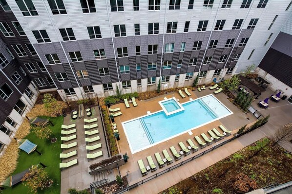A rooftop pool with lounge chairs and a small hot tub, surrounded by a garden and a building with a modern design.