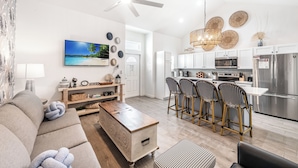 A modern living room with a sofa, coffee table, and a TV. Adjacent is a kitchen with a dining area, featuring a refrigerator, microwave, and bar stools.